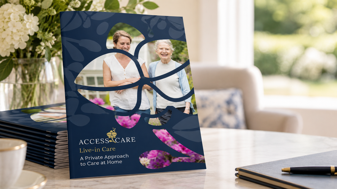 A printed brochure or catalog booklet titled ACCESS CARE stands upright on a tabletop in a real indoor setting with flowers, a chair, and a window visible in the background. A stack of similar booklets lies beside it, and a pen rests on a notebook in the foreground.
