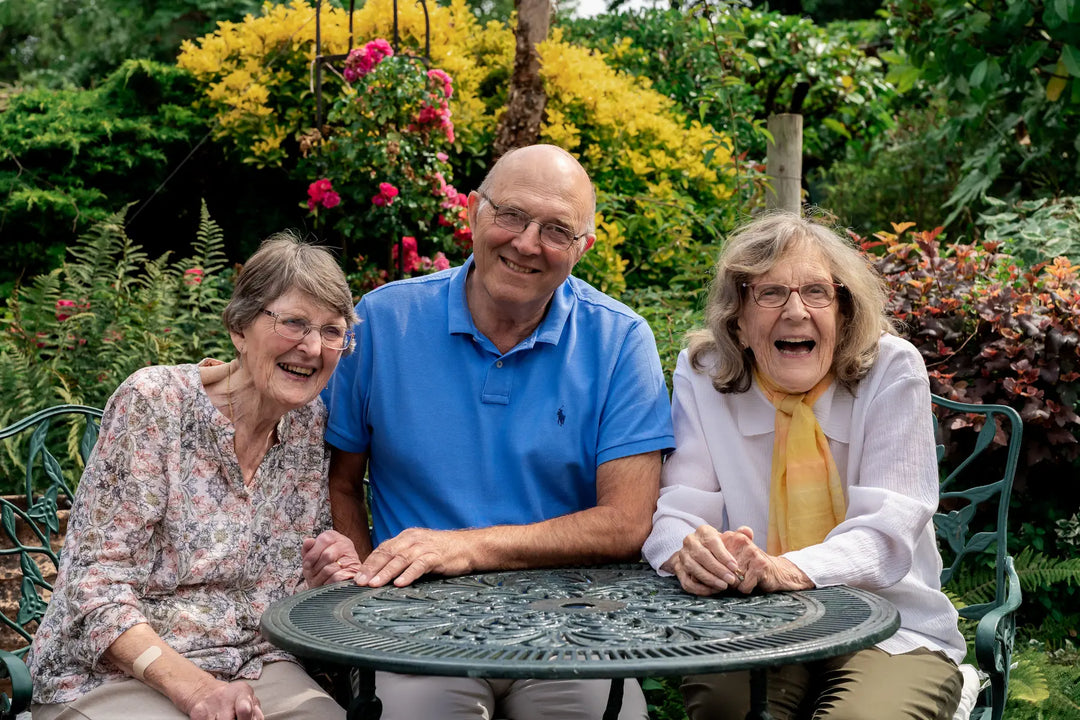 Three smiling older adults sitting together at an outdoor round table in a garden setting, with greenery and flowering plants in the background.