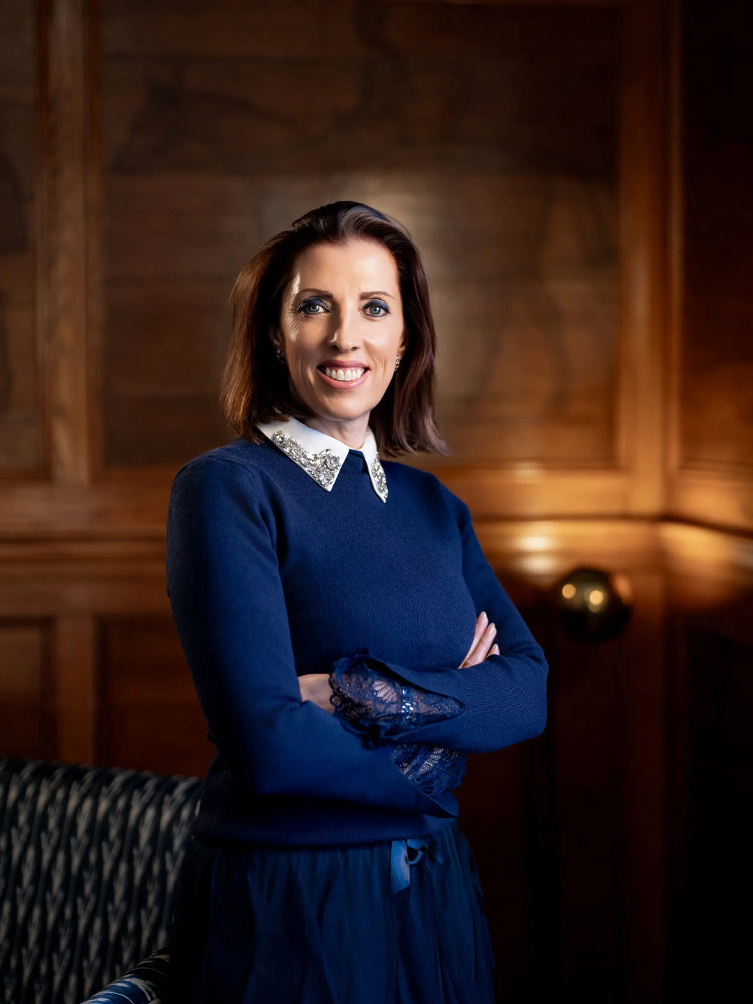 A woman with short brown hair stands with arms crossed, wearing a navy blue sweater over a dark dress with a white lace collar, set against a warm wood-paneled background. The woman is Tiggy Bradshaw CEO of Access Care