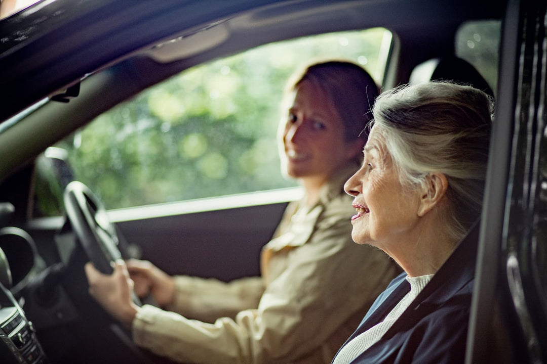 An older woman sits in the passenger seat of a car while a younger woman drives, captured in a warm, softly lit interior with blurred greenery outside the window.