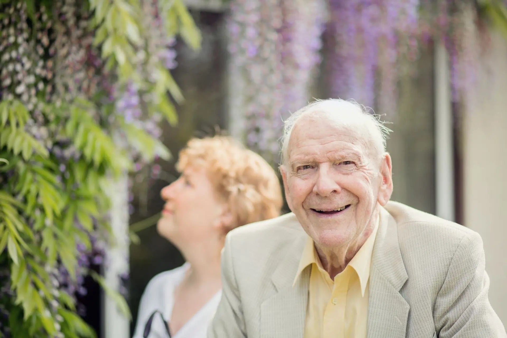Elderly gentleman in garden with live-in carer in background with purple wisteria