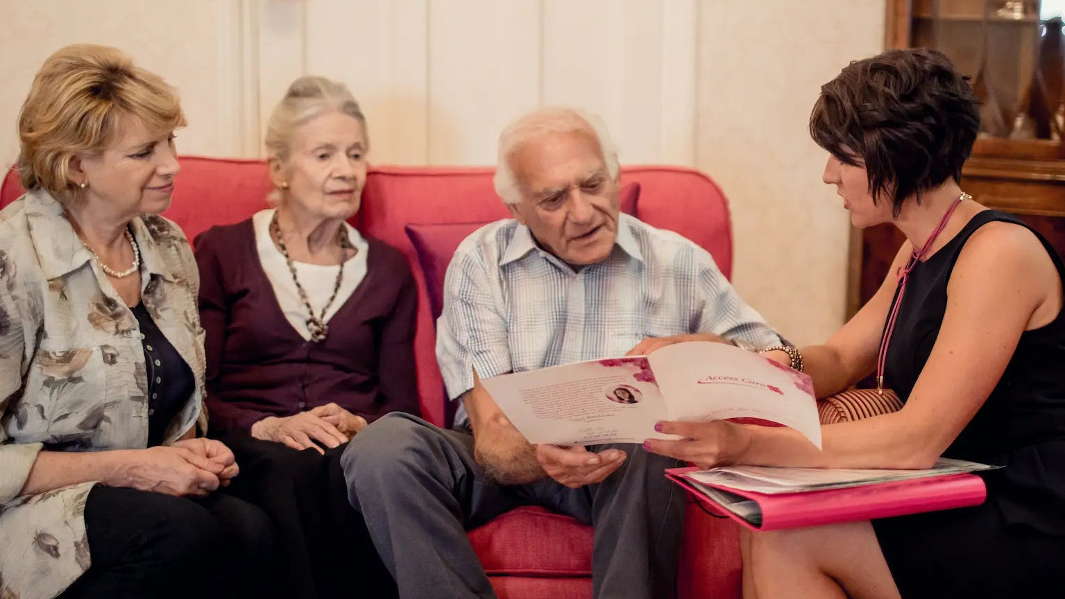 Two older adults sitting on a red couch with two younger women, one of whom is holding a folder. Private care consultation taking place in a client’s home